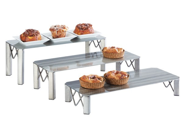 Three metal risers with a polished finish are arranged in a row, each holding pastries. The front riser displays two small round pies, the middle holds two larger tarts, and the back riser showcases four assorted pastries.