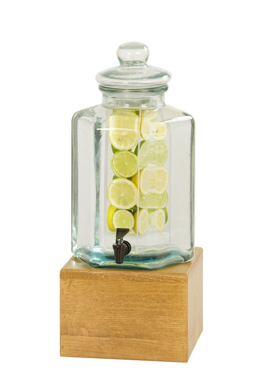 The Madera Beverage Dispenser is a clear glass dispenser with a lid, resting on a wooden base. Filled with water and citrus slices, it features an easy-pour black spigot. The plain white background highlights its elegant design.