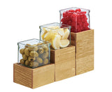Three glass jars sit on a tiered Madera Single Jar Riser made of rustic pine. The jars hold green olives, lemon wedges, and red cherries, from left to right and bottom to top, each fitting neatly atop a different level of the wooden blocks.