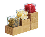 Three glass jars sit on a tiered Madera Single Jar Riser made of rustic pine. The jars hold green olives, lemon wedges, and red cherries, from left to right and bottom to top, each fitting neatly atop a different level of the wooden blocks.