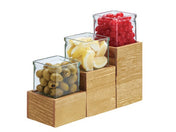Three square glass jars on the rustic pine Madera Single Jar Riser hold green olives, lemon wedges, and red cherries. The tiered wooden blocks create stepped heights, neatly displaying each filled jar against a white background.