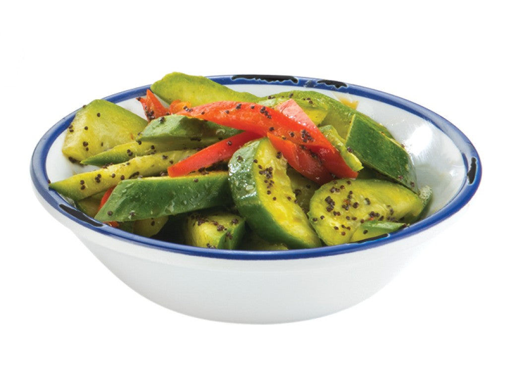A white melamine bowl with a blue rim is filled with sliced cucumbers and red bell peppers, sprinkled with black mustard seeds. The fresh, glossy vegetables suggest a light dressing. The background is plain white.