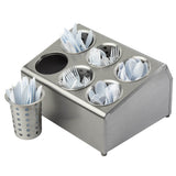 A stainless steel cutlery dispenser with separate compartments holds white plastic forks, spoons, and knives. Beside it, a flatware cylinder contains more utensils. The background is plain and uncluttered.