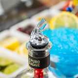 A BPA free, premium liquor pourer is inserted in a bottle labeled “100% Puro Agave.” In the background are cocktail garnishes—lemon slices, lime wedges, cherries, and a blue cocktail with a lemon wheel.