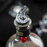 A close-up of a bottle with a BPA free premium liquor pourer inserted in the neck. The label reads “PURO AGAVE.” The background is dark and out of focus, with hints of glassware and textured surfaces.