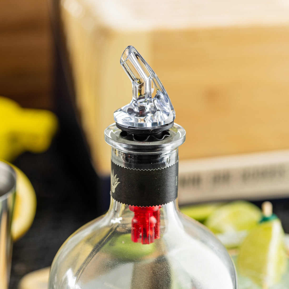 A close-up of a bottle with a BPA free, premium liquor pourer attached to its neck. The bottle cap area is sealed with black and red packaging. In the blurred background, lime wedges and part of a wooden box are visible.