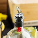 A close-up of a bottle with a BPA free, premium liquor pourer attached to its neck. The bottle cap area is sealed with black and red packaging. In the blurred background, lime wedges and part of a wooden box are visible.