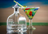 A clear, BPA free, empty bottle of Gran Patrón Platinum tequila sits in the foreground. Behind it is a colorful margarita glass with a salted rim and lime garnish, set against a blurred beach-themed background.