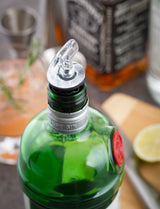 A close-up of a green gin bottle with a BPA free Proper Pour Widemouth Pourer attached. In the blurred background, there’s a cocktail with rosemary, sliced lime, a jigger, and another premium liquor bottle, suggesting a drink-making scene.