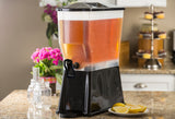 A large, clear 3 gallon beverage dispenser filled with iced tea sits on a kitchen counter. Featuring a black base and spigot, this NSF approved Slimline Beverage Dispenser is surrounded by lemon wedges, mason jars, pink flowers, and cupcakes.