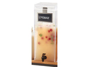 A clear 3 gallon beverage dispenser labeled Lemonade contains a light yellow beverage with red and green fruit pieces floating inside. The dispenser has a small black spigot near the bottom for pouring drinks.