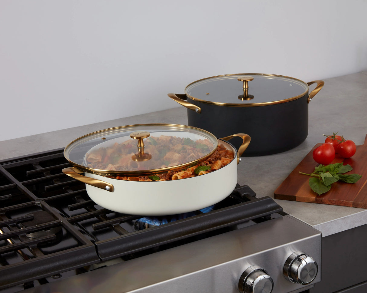 A stovetop features a shallow white pot with a Gold Accent Glass Cover, holding food, while a black pot sits nearby. A cutting board with tomatoes and basil rests beside the pots. The kitchen showcases a sleek, modern Spring USA design.