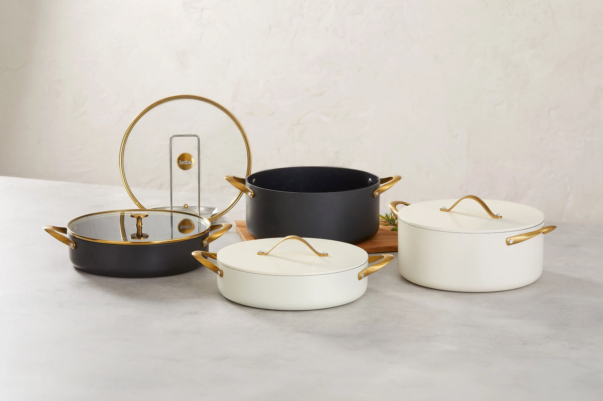 Four elegant Spring USA pots with gold handles and lids are displayed on a light surface. Two are black Buffet Chafer Dishes with Gold Accent Glass Covers, and two are white with solid lids. A sprig of herbs rests near a pot on a wooden board against a neutral background.