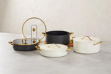 Four elegant Spring USA pots with gold handles and lids are displayed on a light surface. Two are black Buffet Chafer Dishes with Gold Accent Glass Covers, and two are white with solid lids. A sprig of herbs rests near a pot on a wooden board against a neutral background.