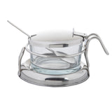 A clear glass container sits in a round metal holder with a handle. A metal lid covers the bowl, featuring a notch for a metal spoon. The holder has a wide, flat base for stability and is part of a stylish condiment jar set.