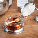 A glass container from a condiment jar set with a metal lid and holder sits on a wooden surface. Inside the honey rests a wooden honey dipper. A sign in the blurred background reads Steel-Cut Oatmeal, near other shiny metal containers.