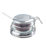 A small glass container filled with dried red chili flakes sits in a shiny metal holder with a lid and curved handle. A metal spoon rests inside, and the dishwasher safe condiment jar set is arranged on a matching round tray against a white background.