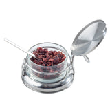 A glass container filled with dried cranberries sits in a shiny silver metal holder. The holder has a round base, an open hinged lid, and a small silver spoon inside. This stylish condiment jar set is perfect for the table and is dishwasher safe.