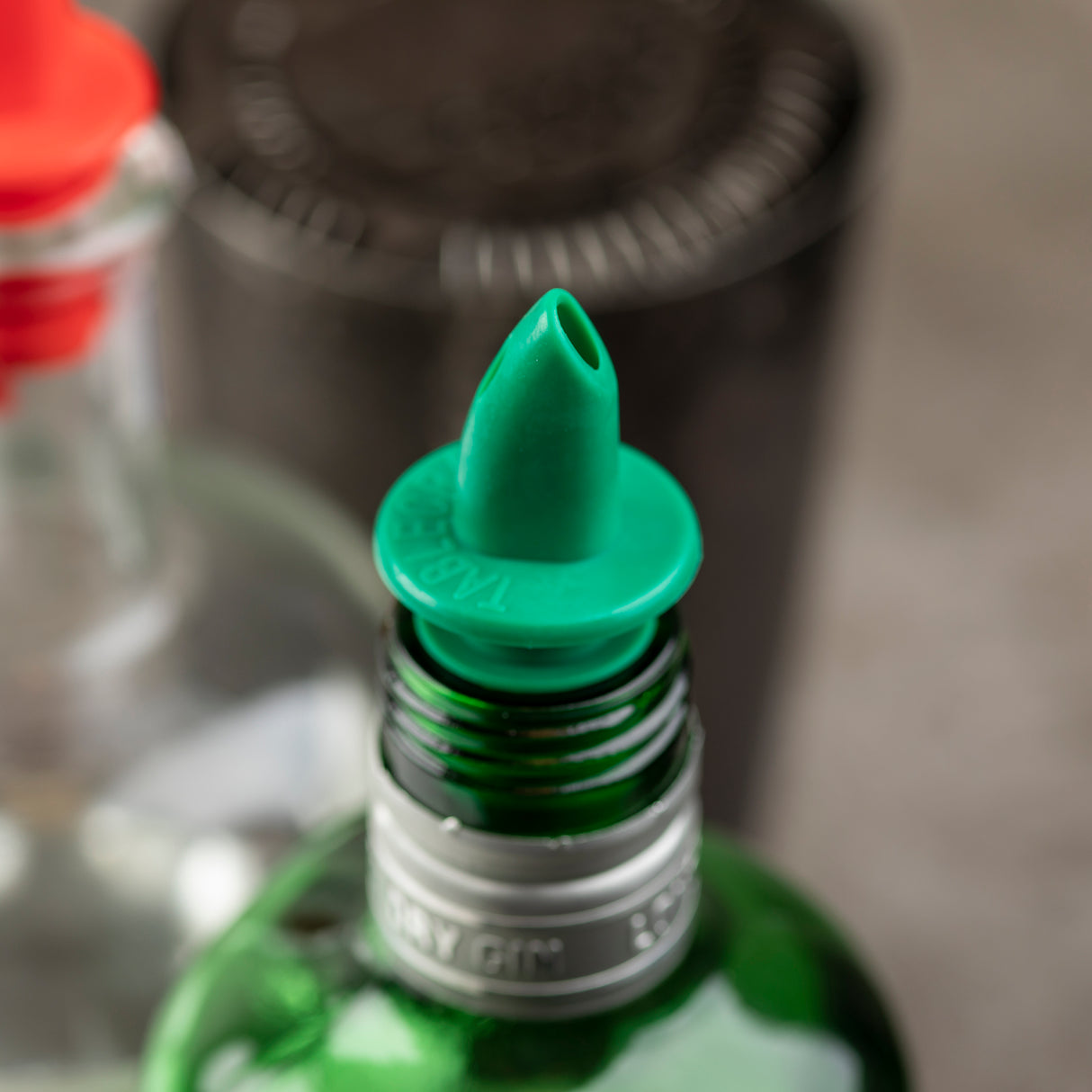 A close-up of a green liquor bottle with a TableCraft Free Flow Pourer inserted in the neck. Other out-of-focus bottles with red caps are in the background, emphasizing the pourer’s spout and bottle rim.