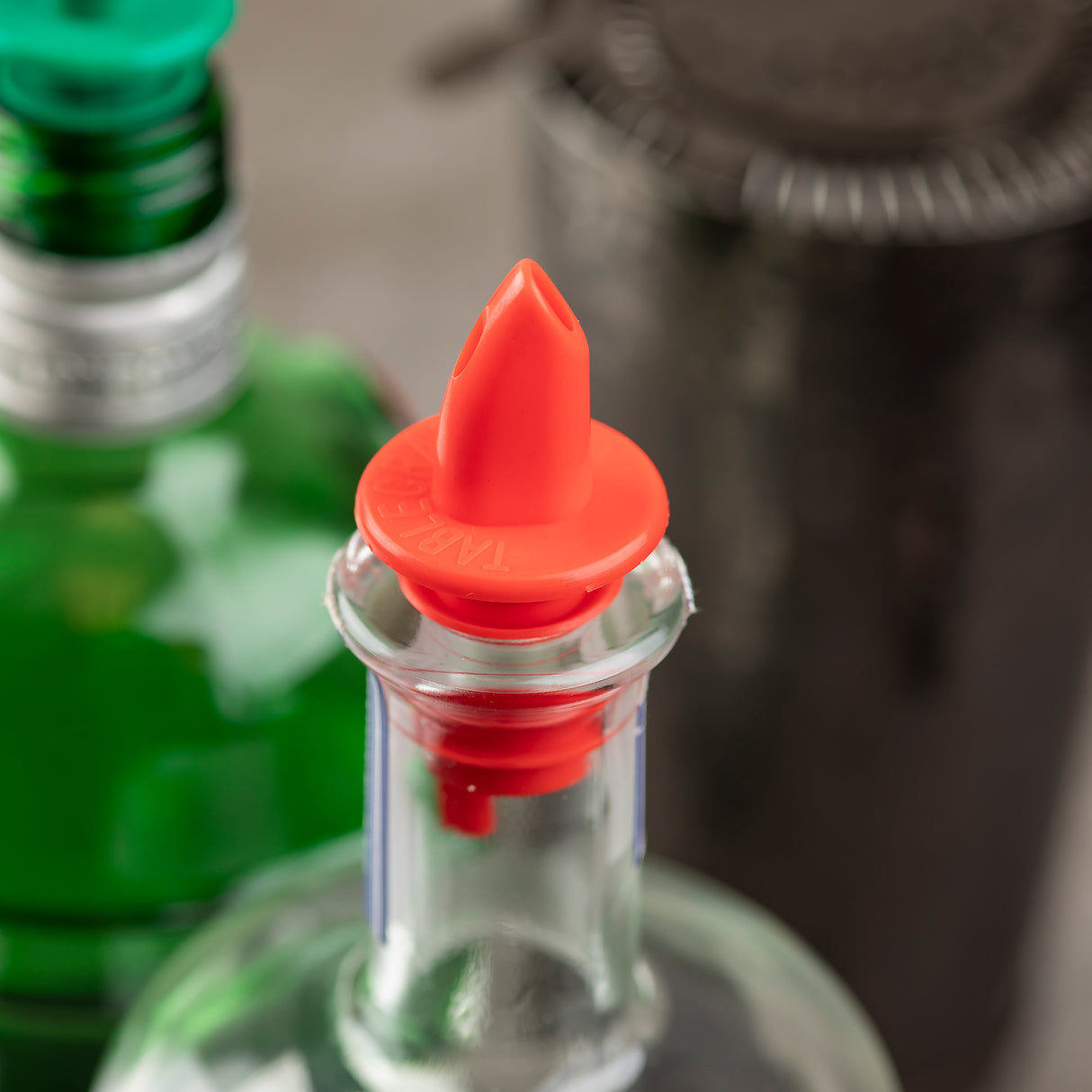 A close-up of a glass bottle with a bright red TableCraft Products Free Flow Pourer inserted in its neck. A green bottle with a green cap and a dark, out-of-focus object are visible in the blurred background, suggesting a bar or kitchen setting.