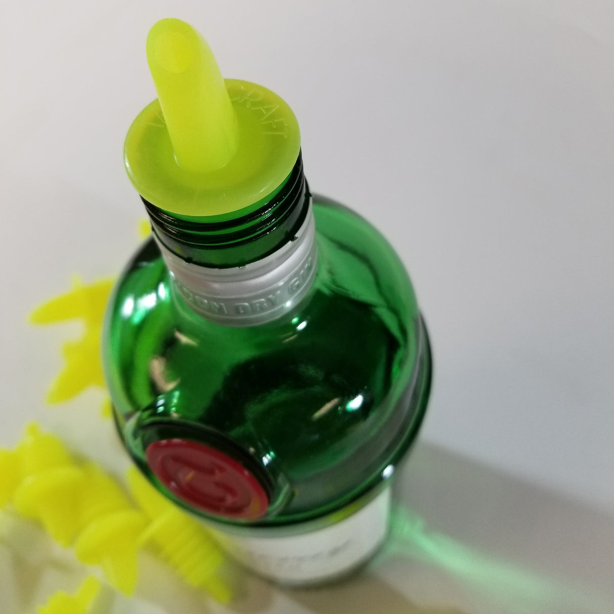 A green glass bottle with a yellow TableCraft Economy Free Flow Pourer inserted into its neck is shown from above. Several additional yellow spouts are visible lying on the white surface next to the bottle, which has a red seal near the top.