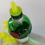 A green glass bottle with a yellow TableCraft Economy Free Flow Pourer inserted into its neck is shown from above. Several additional yellow spouts are visible lying on the white surface next to the bottle, which has a red seal near the top.