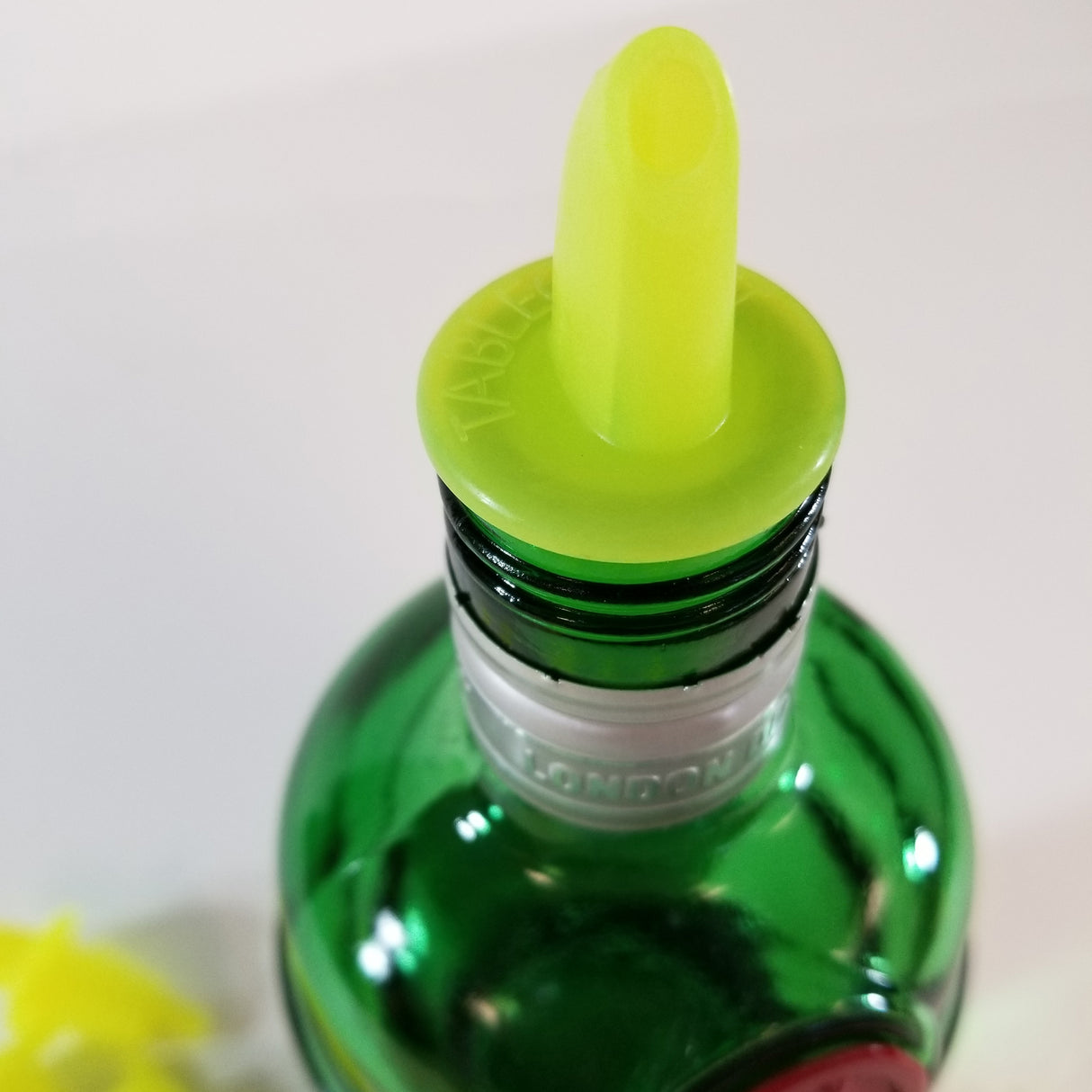 A close-up of the top of a green glass bottle with a metallic neck ring labeled LONDON DRY. The bottle is fitted with a bright yellow TableCraft Economy Pourer. The background is plain and out of focus.
