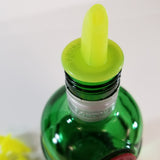 A close-up of the top of a green glass bottle with a metallic neck ring labeled LONDON DRY. The bottle is fitted with a bright yellow TableCraft Economy Pourer. The background is plain and out of focus.