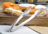 A close-up of a tray filled with donuts topped with orange and white icing. In front, a stainless steel Dalton II Collection Tong Server from TableCraft Products rests on the edge, with a white tablecloth and beige napkin partially visible underneath.