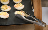 A close-up of deviled eggs arranged on a black tray, sprinkled with herbs and paprika. A Dalton II Collection Tong Server by TableCraft Products rests on the tray’s edge, set atop a wooden display stand amid an out-of-focus buffet setting.