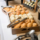 Wooden trays filled with assorted bread rolls and pastries are arranged on a white tablecloth. A stainless steel tong server from the Dalton II Collection rests atop the trays. Spoons and forks are nearby, with deviled eggs in the background.
