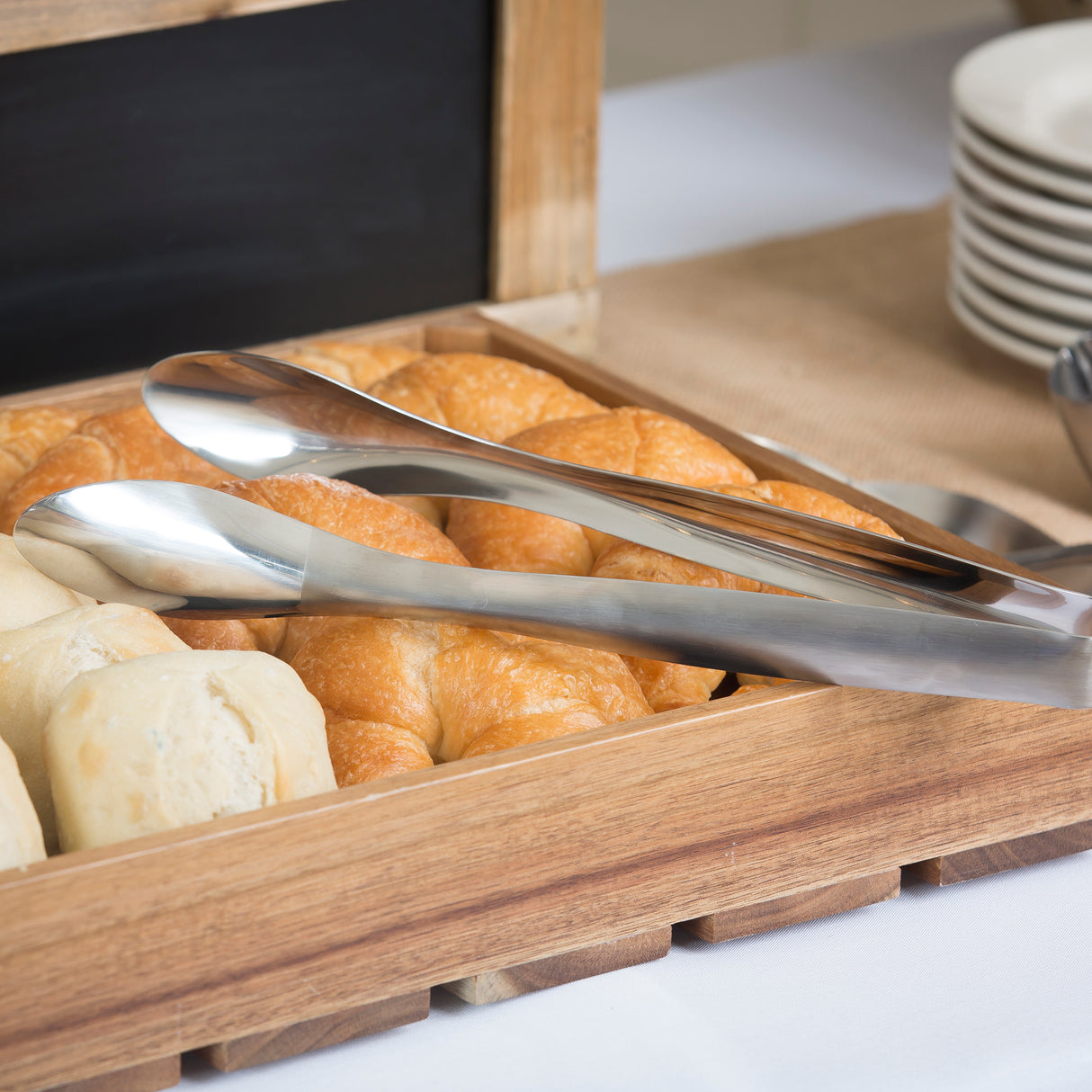 A wooden tray holds golden croissants and bread rolls. A pair of stainless steel tong server from the Dalton II Collection rests atop the pastries. In the background, stacked white plates and part of a chalkboard suggest a buffet setting.