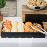 A black tray from the Dalton II Collection on a white tablecloth holds cheese rolls, plain rolls, and croissants. Stainless steel tong server rests on the tray. Behind, a bowl of rice salad sits on a wooden riser with a potted plant and stone wall behind.