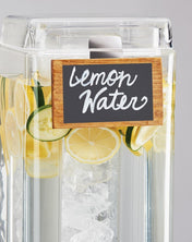 A clear beverage dispenser filled with ice, lemon slices, and cucumber slices features a chalkboard beverage sign reading "lemon water" in white cursive. The dispenser is set against a plain, light gray background.