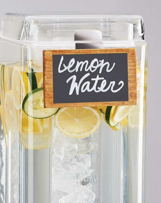 A clear beverage dispenser filled with ice, lemon slices, and cucumber slices features a chalkboard beverage sign reading "lemon water" in white cursive. The dispenser is set against a plain, light gray background.