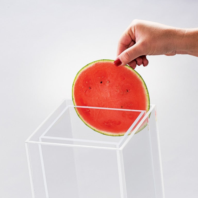 A hand with red-painted nails is placing a round slice of watermelon into a transparent, BPA free rectangular box against a plain light background, perfect for prepping fruit for your 3 gallon beverage dispenser.