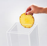 A hand with red nail polish holds a round slice of pineapple above a transparent, empty BPA free acrylic box against a plain light background. The pineapple slice is bright yellow with a slightly rough brown edge.