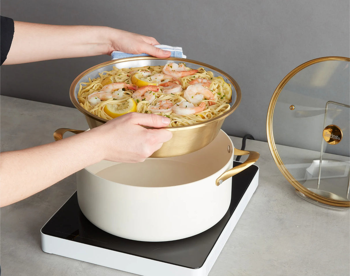 A person holds a pan of cooked pasta with shrimp, lemon slices, and herbs above a white pot on an electric stovetop, preparing to combine or strain the ingredients. Nearby, a buffet chafer dish with a gold accent insert rests on the counter.