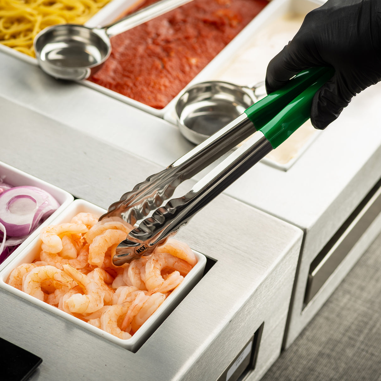 A gloved hand uses 12 inch tongs with a green handle to pick up shrimp from a small white container. Nearby are containers with sliced red onions, pasta noodles, tomato sauce, and another white sauce in a buffet-style setup. Dishwasher safe tongs.