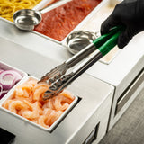 A gloved hand uses 12 inch tongs with a green handle to pick up shrimp from a small white container. Nearby are containers with sliced red onions, pasta noodles, tomato sauce, and another white sauce in a buffet-style setup. Dishwasher safe tongs.