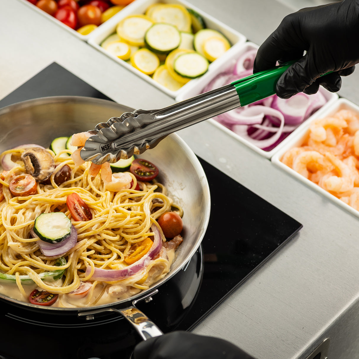 A chef wearing black gloves uses 12 inch tongs to add shrimp to a skillet of creamy pasta with cherry tomatoes, zucchini, mushrooms, and red onion. Containers of sliced vegetables and shrimp sit on the prep counter; the tongs are dishwasher safe.