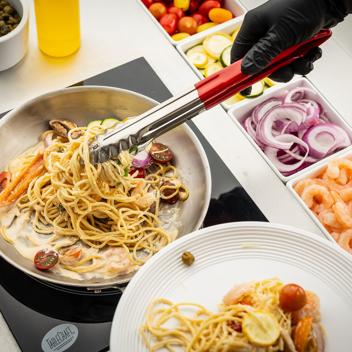 A gloved hand uses spring steel tongs to serve creamy pasta with shrimp, cherry tomatoes, and vegetables from a skillet onto a white plate. Nearby, containers hold sliced onions, shrimp, tomatoes, and zucchini on a white countertop.