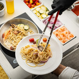 A gloved hand uses spring steel tongs to serve creamy spaghetti with shrimp, vegetables, and cherry tomatoes onto a white plate. Below is a pan on the stovetop, with toppings like zucchini and onions arranged in containers nearby.