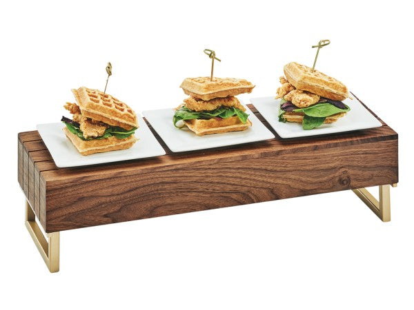 Three waffle sandwiches with chicken and greens are placed on white square plates atop a mid-century reversible riser with brass frame; each sandwich is held together with a decorative skewer.