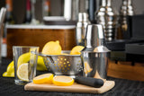 A stainless steel bar shaker, an empty glass with a lemon slice, halved lemons on a cutting board, and a muddler sit on the counter. Behind them is a metal colander with whole lemons, while bar tools and bottles blur softly in the background.