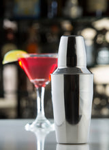 A shiny 16 oz stainless steel bar shaker sits in the foreground. In the blurred background, a red cocktail in a martini glass is garnished with a lemon slice, set against bottles visible at the bar.