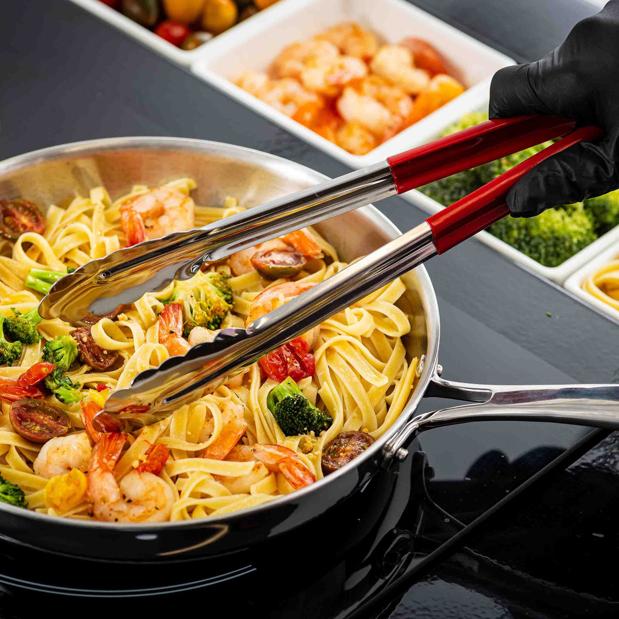 A hand wearing a black glove uses red-handled, spring steel tongs to mix fettuccine pasta with shrimp, broccoli, and tomatoes in a stainless steel pan on the stovetop. In the background are divided trays with shrimp, broccoli, and other ingredients.