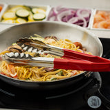 A close-up of a frying pan with creamy pasta, shrimp, and vegetables on a stove. Red-handled vinyl-coated tongs rest on the pan’s edge. In the background, trays hold sliced zucchini, onions, and shrimp, slightly out of focus.