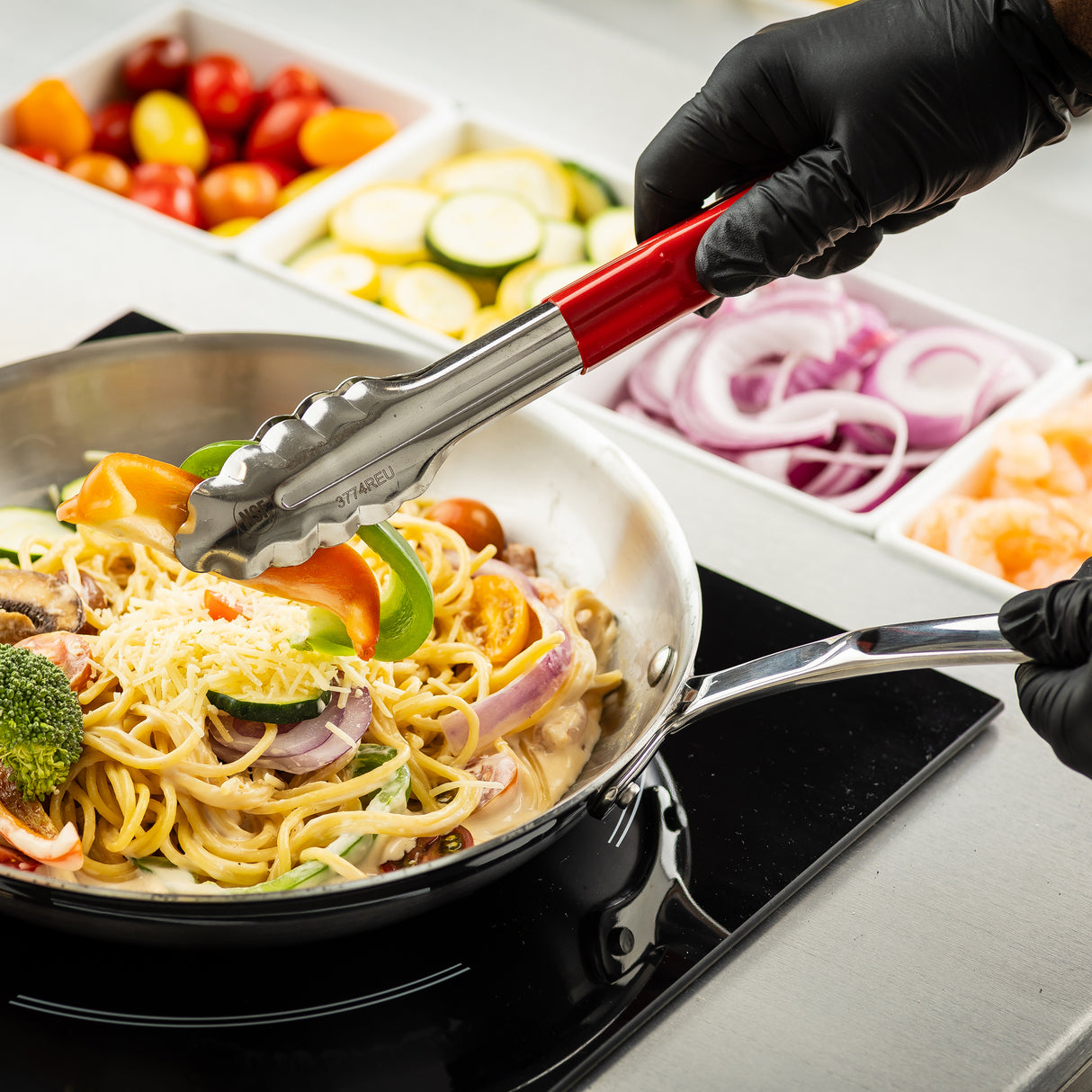 A person wearing black gloves uses spring steel tongs to prepare creamy pasta with vegetables and cheese in a pan. Beside the pan are containers of sliced onions, shrimp, zucchini, and cherry tomatoes, showing fresh ingredients for cooking.