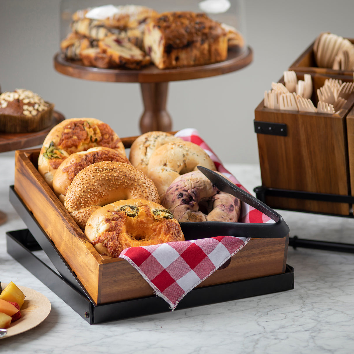 A wooden tray lined with red and white checkered cloth holds assorted bagels, including sesame and cheese-topped varieties, with stainless steel tongs resting on the tray. In the background are pastries on a cake stand and a wooden box with utensils.